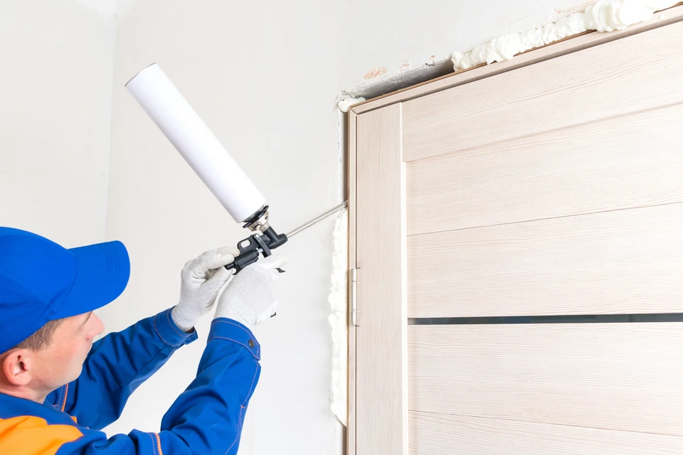 un maître en uniforme de protection bleu installe une porte intérieure, éliminant l'espace avec la mousse de montage