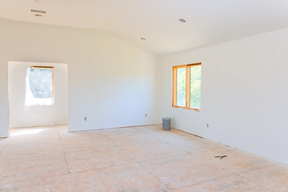 Ouvrier en salopette bleue et casque blanc applique de l'enduit sur un mur intérieur avec une spatule, un seau de pâte à ses pieds